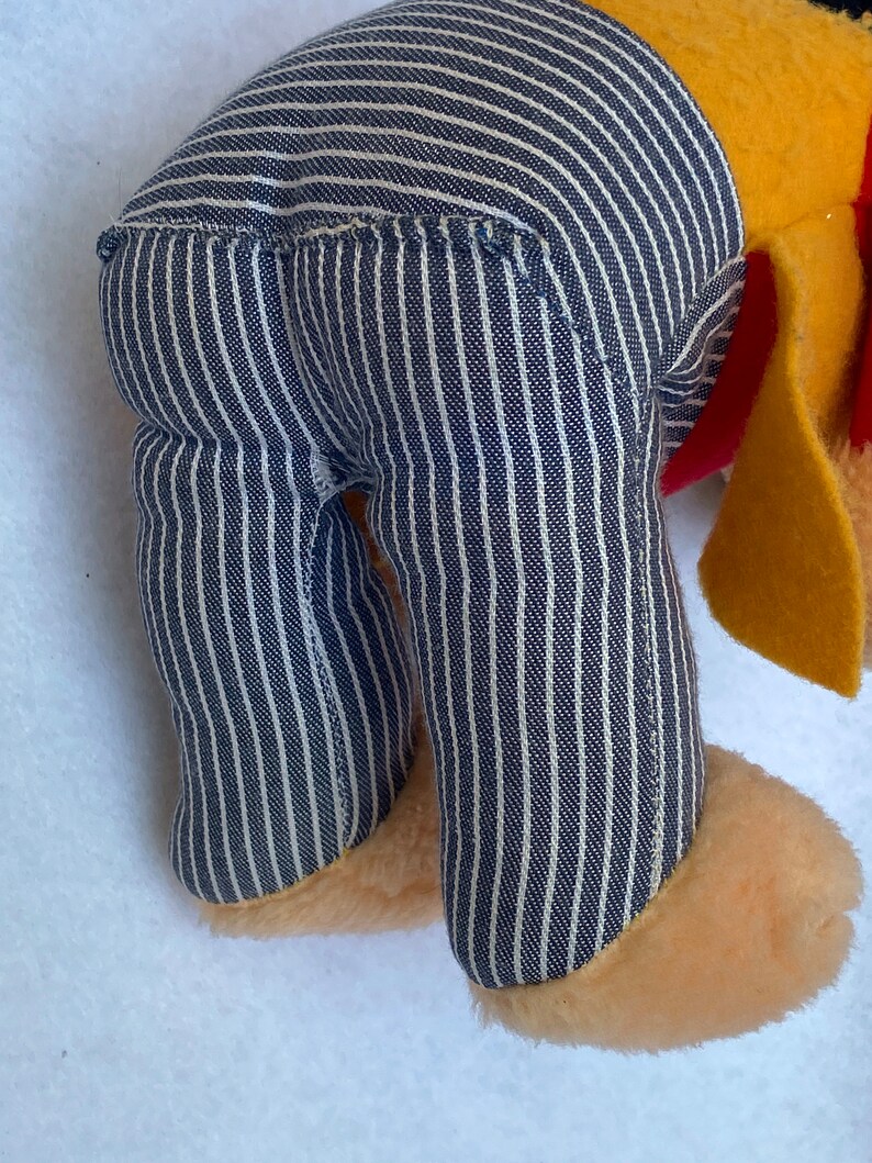 May include: A plush toy with a yellow body, blue and white striped pants, and a red collar. The toy is standing on a white surface.