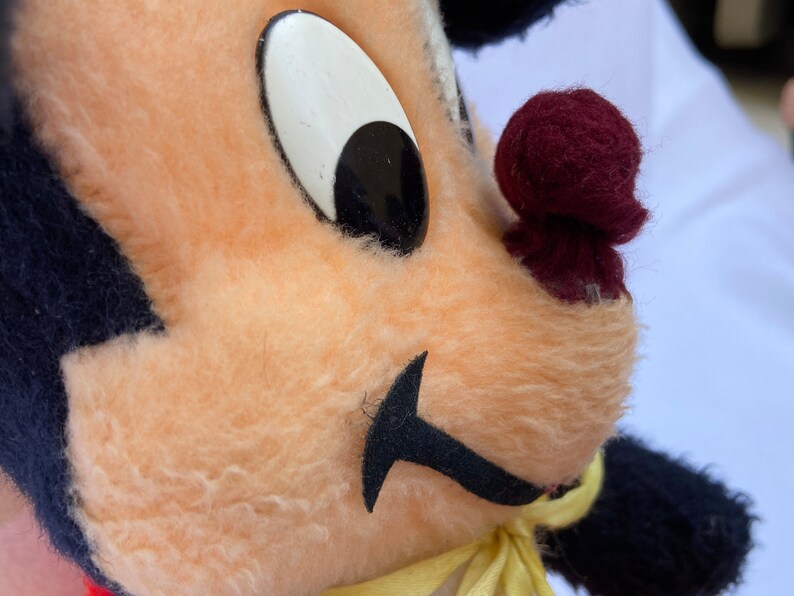 May include: Close-up of a plush toy with black fur, a peach-colored face, a black mustache, a large white eye with a black pupil, and a maroon nose.
