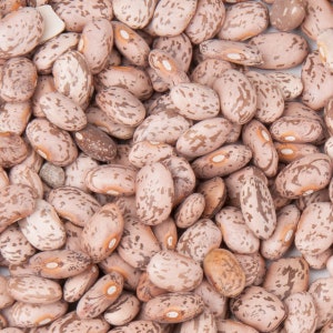 Puede incluir: Primer plano de un montón de frijoles pintos. Los frijoles son ovalados, con un color base beige claro y marcas moteadas marrones. Algunos frijoles tienen un color marrón más oscuro. Los frijoles están secos y listos para cocinar.