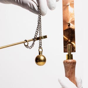 May include: A close-up of two objects held by gloved hands against a white background. One is a brass balance scale with a chain and a spherical weight. The other is a copper-colored metal object with a wooden handle.