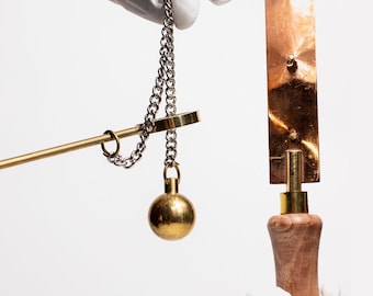 May include: A close-up of two objects held by gloved hands against a white background. One is a brass balance scale with a chain and a spherical weight. The other is a copper-colored metal object with a wooden handle.