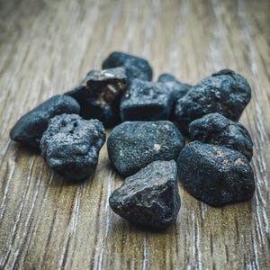 Könnte beinhalten: Nahaufnahme eines Haufens dunkler, unregelmäßig geformter Steine. Die Steine sind tiefschwarzgrau, einige weisen braune Farbtöne auf. Die Textur wirkt rau und natürlich, mit unterschiedlichen Größen und Formen. Die Steine liegen auf einer hellbraunen, strukturierten Oberfläche.