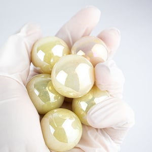 Puede incluir: Un puñado de canicas de vidrio iridiscentes en tonos amarillos y rosa pálido. Las canicas tienen una superficie brillante y reflectante y se sostienen en una mano enguantada sobre un fondo blanco. Estas canicas decorativas son ideales para manualidades o decoración del hogar.