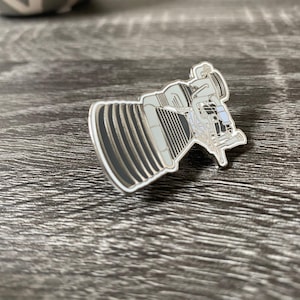 May include: A silver-colored enamel pin depicting a detailed illustration of a rocket engine. The pin features a ribbed, cylindrical section and intricate mechanical details in shades of gray and black. The pin is set against a wooden surface.