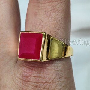 以下が含まれることがあります： 四角い赤い宝石がついた金の指輪。指にはめられています。