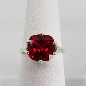 May include: A silver ring featuring a large, red, cushion-cut gemstone.