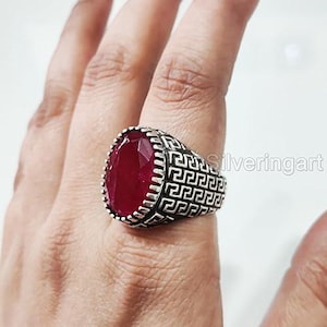 May include: A silver ring with a large, oval-cut, deep red gemstone. The ring has a detailed, geometric design around the band. The gemstone is the focal point, catching the light and adding a bold statement to the jewelry.