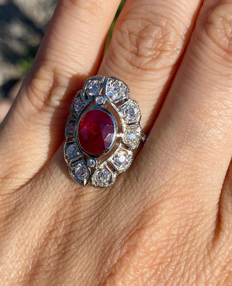 May include: A silver ring with a large red ruby surrounded by smaller diamonds. The ring is set in a floral design.