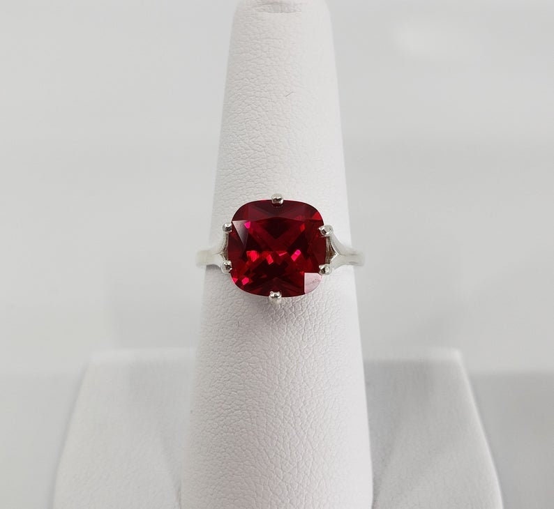 May include: A silver ring featuring a large, red, cushion-cut gemstone.
