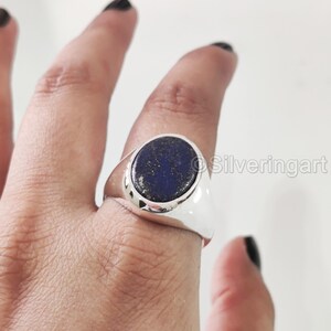 Può includere: Un anello d'argento con una grande pietra ovale blu scuro. L'anello ha una finitura lucida e brillante. La pietra ha un aspetto macchiettato. L'anello è su un dito.