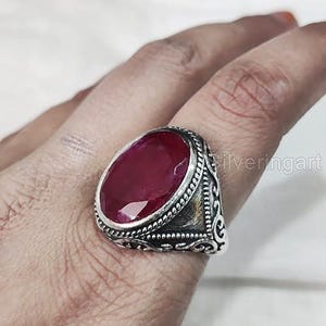 May include: A large, oval ruby gemstone ring. The dark red gemstone is set in a silver-toned, ornate band. The band has a detailed, textured design. The ring is shown on a finger.