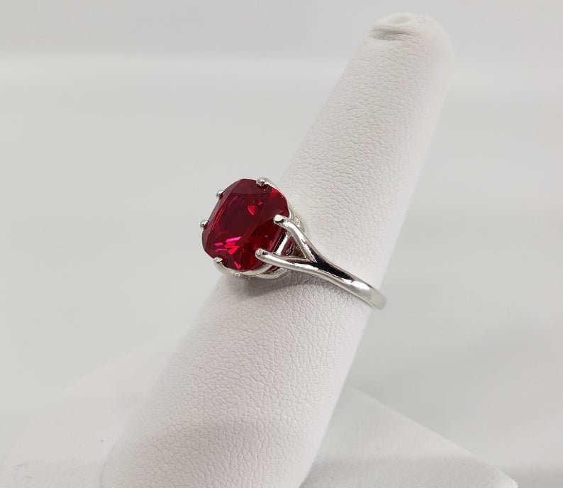 May include: A silver ring featuring a large, red, cushion-cut gemstone.
