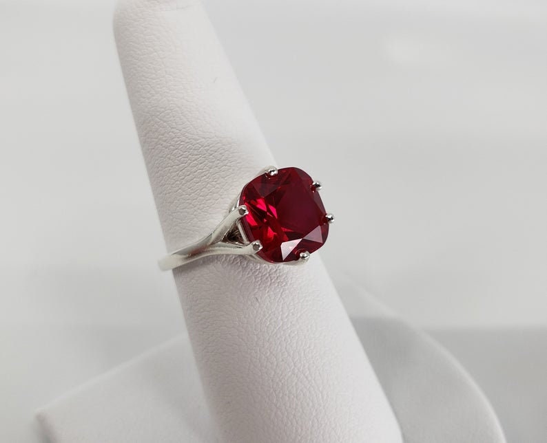 May include: A silver ring featuring a large, red, cushion-cut gemstone.