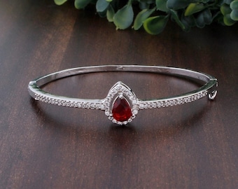 Puede incluir: Un brazalete de plata en forma de bangle con una piedra roja en forma de lágrima rodeada de piedras de circonita cúbica transparentes.