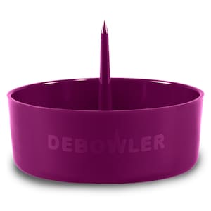May include: A magenta-colored Debowler tool with a cylindrical base and a central spike. The word "DEBOWL" is embossed on the side. The tool is designed for cleaning and preparing smoking devices.