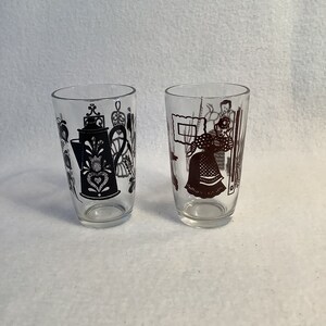 Op de afbeelding: Twee heldere drinkglazen met decoratieve zwarte en bruine ontwerpen. Eén glas heeft een koffiepot en bloemmotieven, terwijl de andere figuren in traditionele klederdracht afbeeldt. De glazen zijn cilindrisch en lijken vintage.