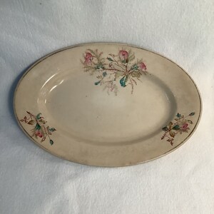 以下が含まれることがあります： 金色の縁取りが付いた、楕円形のクリーム色のサービング皿。ピンクと緑の花柄模様が施されています。花柄のデザインは中央と側面にあります。この皿はおそらくセラミック製です。