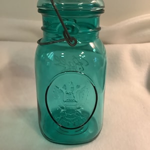 Puede incluir: Un tarro de vidrio de color turquesa con asa y tapa de alambre metálico. El tarro presenta un emblema de águila en relieve y la palabra "SIDE" en la tapa. Estilo vintage, para conservas o almacenamiento.