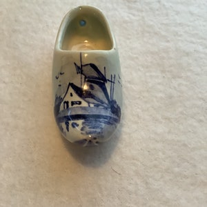 May include: A small, ceramic Dutch clog painted with a blue and white landscape scene. The miniature shoe features a detailed depiction of a windmill and houses, typical of Dutch imagery. The clog is a decorative item, likely a souvenir.