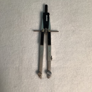 Pode incluir: Um compasso prateado e preto com a marca "charvoz". A ferramenta tem duas pernas ajustáveis, uma com ponta e outra para lápis. Projetado para desenhar círculos e arcos, comumente usado em desenho técnico.