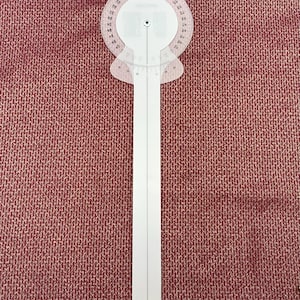 Puede incluir: Un transportador de plástico blanco con una escala de grados circular transparente y un brazo largo y recto. El transportador está diseñado para medir ángulos y se encuentra sobre un fondo texturizado de color marrón rojizo.