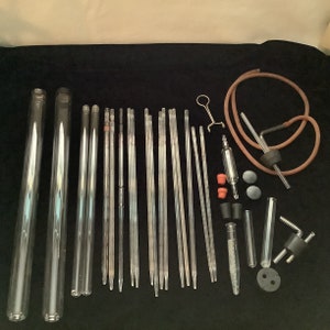 May include: A collection of clear glass laboratory equipment, including test tubes, pipettes, and flasks. The assortment features various sizes and shapes, with some pieces connected by rubber tubing and stoppers. The items are arranged on a dark surface.