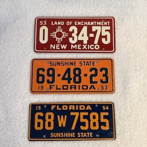 Puede incluir: Tres placas de matrícula antiguas. La placa superior es roja con letras blancas y dice "NEW MEXICO" y "LAND OF ENCHANTMENT". La placa del medio es naranja con letras azul oscuro y dice "FLORIDA" y "SUNSHINE STATE". La placa inferior es azul oscuro con letras naranjas.