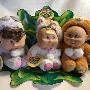 May include: Three Cabbage Patch Kids Snuggles dolls are presented on a green leaf-shaped stand. The dolls are dressed in animal-themed outfits: a grey hamster, a white bunny, and a brown lion. Each doll has a tag that reads "Cabbage Patch Kids Snuggles".