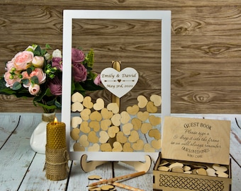 Könnte beinhalten: Weißer Holzrahmen mit einem herzförmigen Schild, auf dem "Emily & David May 3rd, 2020" steht. Der Rahmen ist mit kleinen Holzh Herzen gefüllt. Neben dem Rahmen befindet sich eine Holzkiste mit einem Schild, auf dem "Gästebuch Bitte unterschreiben Sie ein & legen Sie es in den Rahmen, damit wir uns immer an unsere Lieben erinnern, die gekommen sind" steht.