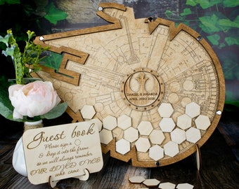 May include: A wooden guest book shaped like the Millennium Falcon from Star Wars. The ship is engraved with a blueprint design and has a small circle in the center with the text "DANIEL & AMANDA APRIL 23RD 2020". The guest book comes with small wooden hexagonal pieces that guests can sign and place on the ship.