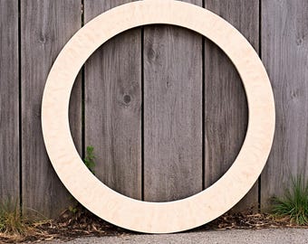 May include: A large, unfinished wooden circle frame, resting against a weathered gray wooden fence. The frame is made of light-colored wood and has a smooth, rounded edge. The background includes a concrete surface and some greenery.