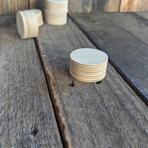 May include: Several stacks of small, round, unfinished wooden discs are arranged on a weathered wooden surface. The discs are light brown and appear to be made of layered wood. The background is a vertical wooden panel.
