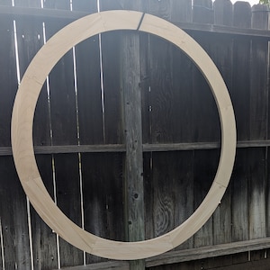 May include: A large, unfinished wooden ring, likely for crafting or decoration. The ring is made of light-colored wood and is composed of several interlocking pieces. It is positioned against a weathered wooden fence.