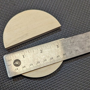 May include: Two unfinished wooden half-circle shapes and a metal ruler. The ruler is marked in inches, with the half-circle measuring approximately 3 inches in diameter. The wood has a light, natural color.