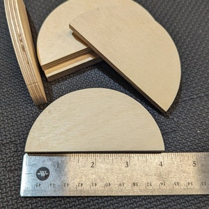 May include: Several unfinished, semi-circular wooden pieces are arranged on a dark surface. A metal ruler shows the pieces are approximately 4 inches in diameter. The wood has a light, natural color and visible grain.