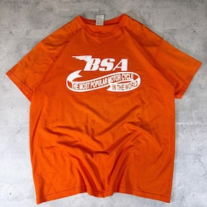 May include: Orange t-shirt with the white text "BSA THE MOST POPULAR MOTOR CYCLE IN THE WORLD". The shirt has short sleeves and a crew neck. The BSA logo features a wing design.