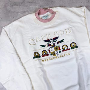 May include: Cream-colored sweatshirt with a pink collar and the embroidered text "CAPE COD" in gold. Below the text is a colorful emblem with a lighthouse, eagle, and other nautical symbols. The text "MASSACHUSETTS" is embroidered below the emblem.