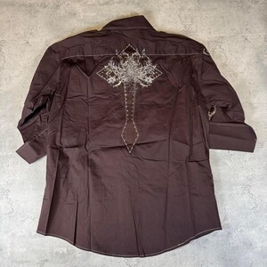 May include: A brown long-sleeve button-down shirt with a white embroidered design on the back. The design features a cross with a floral pattern.