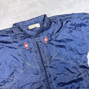 May include: A navy blue vintage jacket with a full zipper and embroidered geometric designs in red, turquoise, and yellow. The jacket has white stitching and a collar.