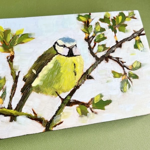 May include: Oil painting of a blue tit bird perched on a branch with green leaves. The bird has a yellow breast and a blue head. The artwork is on a white canvas with a green background. The artist's initials, JZK, are in the lower right corner.