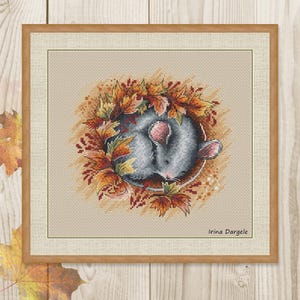 Può includere: Opera d'arte a punto croce di un topo addormentato circondato da una ghirlanda di foglie autunnali nei toni dell'arancione, giallo e marrone. Il topo è grigio con orecchie rosa. L'opera è in una cornice di legno. Il nome dell'artista, Irina Dargele, è in basso a destra.