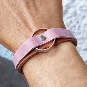 Op de afbeelding: Een lederen armband met een lichtpaarse band over een basis van bruin leer. De twee lagen worden bij elkaar gehouden door een zilverkleurige metalen ring en een kleine zilverkleurige metalen klinknagel voor een eenvoudig ontwerp.
