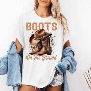May include: White t-shirt featuring a brown cowboy hat and boots graphic with the text "BOOTS On The Ground". The design includes a decorative background. The shirt is a casual wear item.