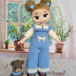 May include: A handcrafted crochet doll wearing a blue jumpsuit, alongside a small brown teddy bear. The doll features blonde hair in two buns adorned with blue flower accents. The jumpsuit has a white bow and lace trim. The teddy bear wears a blue collar. The background shows a window and plants.