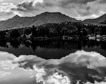 May include: A black and white photograph of a mountain range reflected in a still lake. The sky is cloudy with a bright spot of light shining through the clouds.