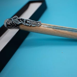 May include: A handcrafted pen with a light wood barrel and a silver-colored metal tip and accents. The pen features a decorative silver clip and is presented in a black box against a blue background.