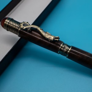 May include: A decorative pen with a brown barrel and ornate metal accents. A detailed dragon figure is featured on the pen's body. The pen has a red gem at the top and a textured tip. The pen is presented on a blue background.
