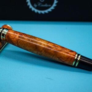 May include: A luxury fountain pen with a wooden barrel and gold-colored accents. The pen is lying on a light blue surface. The pen's cap is off, revealing the nib.
