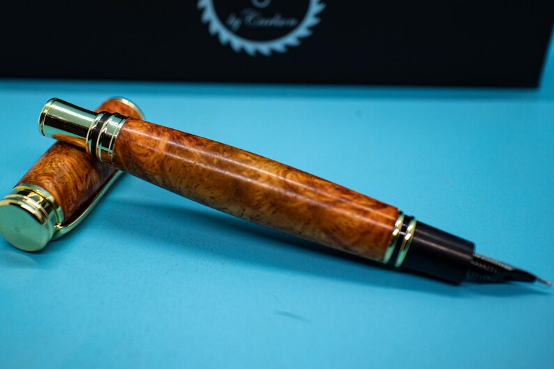 May include: A luxury fountain pen with a wooden barrel and gold-colored accents. The pen is lying on a light blue surface. The pen's cap is off, revealing the nib.