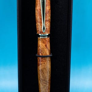 May include: A handcrafted pen with a wood-grain barrel and gold-colored accents, presented in a black box. The pen features a silver clip and a black band. The pen's design showcases a natural wood pattern, adding to its elegant appearance.